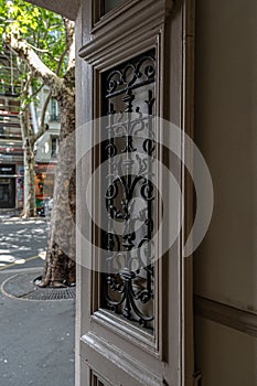 Opened door with ornate antique grating on door window. Blurry background with city street. Beige painted door panel. Architecture