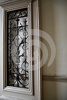 Opened door with ornate antique grating on door window with stone wall background. Beige painted door panel. Architectural details