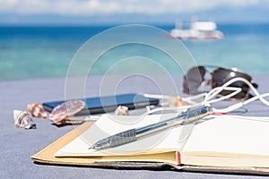 Opened empty notepad is on the table with phone and headphones at the tropical sea background