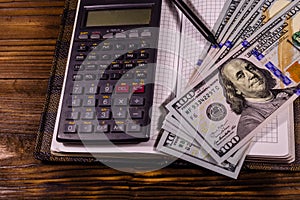 Opened notepad, one hundred dollar banknotes, scientific calculator and pencil on wooden table