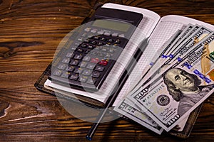 Opened notepad, one hundred dollar banknotes, scientific calculator and pencil on wooden table