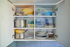 Opened white kitchen cupboard with plates, metal pots and food containers