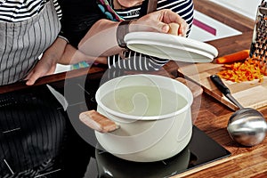 Opened white saucepan with wood pens on the black stove. Cooking a soup - water boiling, check and preparation. Woman`s hand