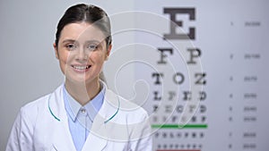 Openhearted lady oculist looking to camera, eyechart on background, profession