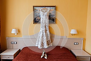 Openwork wedding dress hanging on the picture near the wall next to the bride`s shoes on the bed in the bride`s bedroom