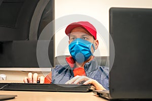 Operator works in the office during the quarantine. A man in a medical mask sits in front of the monitor screens. Specialist works