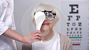 Ophthalmologist closing eye to senior lady in phoropter, checking lens diopter