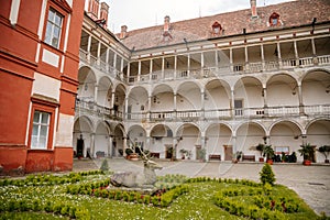 Opocno castle, renaissance chateau, courtyard with arcades and red facade, green lawn with statue and flowers in foreground, sunny