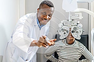 Optician doing optometry eye exam for black african american teen girl patient. Male optometrist with phoropter while examining