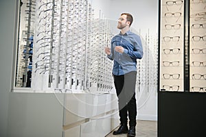 In Optics Shop. Portrait of male client holding and wearing different spectacles, choosing and trying on new glasses at