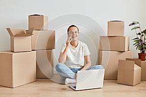 Optimistic beautiful woman wearing white t shirt sitting on floor surrounded with cardboard boxes with belongings and working on
