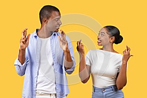 Optimistic black couple with crossed fingers, hoping, yellow background