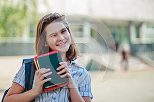 Optimistic Caucasain girl blinks with one eye and smiles. Schoolgirl holds books in hand. Girl with a school backpack photo