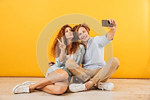 Optimistic couple young man and woman 20s sitting on floor together and showing peace sign while taking selfie on smartphone, iso