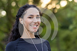 Optimistic diverse sportswoman in black top smiling at camera while listening to motivating playlist in wired earphones