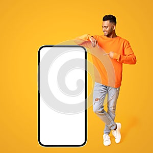 Optimistic happy indian guy posing by huge smartphone with empty blank screen, pointing at display and smiling
