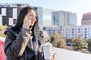 Optimistic Hispanic woman with thermo cup talking on smartphone