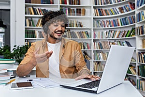 Optimistic indian student using wireless headset and laptop while sitting by desk in library. Confident curly guy