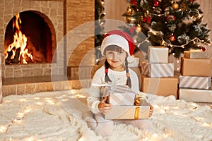 Optimistic little kid wearing white sweater and santa claus hat, sitting on soft carpet with stack of present boxes, posing in