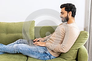 Optimistic man lying on the couch and using laptop computer for messaging, happy businessman working remotely in relaxed