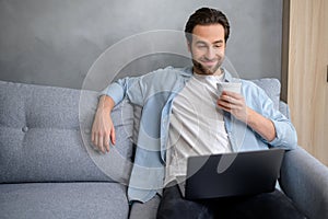 Optimistic man sitting on the couch and using laptop computer. Happy businessman working remotely in relaxed pose