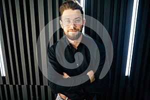 Optimistic Portrait of a Handsome Young Businessman Standing Against modern office Wall While Looking at the Camera with Arms Cros