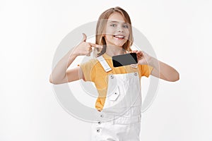Optimistic proud cute blond little girl showing smartphone and pointing mobile screen with pleased enthusiastic