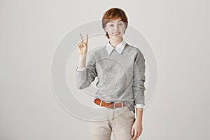 Optimistic redhead female with boyish hairstyle and freckles showing victory or peace gesture, smiling, expressing