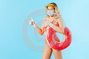 Optimistic woman in swimsuit holding rubber ring, wearing hygienic face mask and gloves to prevent contagious coronavirus