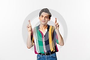 Optimistic young man smiling, holding fingers crossed for good luck, making wish, standing over white background