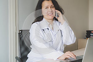 Optomistic doctor woman in medical institution sits at the desktop with computer and speaks on the phone. Phisician is smiling