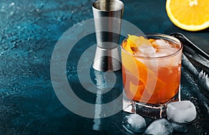 Orange Alcohol cocktail with red vermouth, bitter, soda, orange zest and ice, dark wooden bar counter background, bar tools,