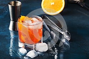 Orange Alcohol cocktail with red vermouth, bitter, soda, orange zest and ice, dark wooden bar counter background, bar tools,