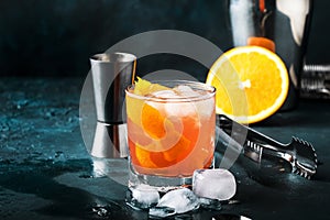 Orange Alcohol cocktail with red vermouth, bitter, soda, orange zest and ice, dark wooden bar counter background, bar tools,