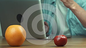 Orange, apple and tomato on table in front of man who using laptop