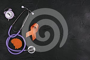 Orange awareness ribbon, brain symbol, stethoscope and alarm clock on a black background. Copy space for text