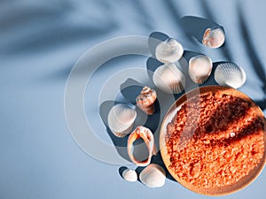 Orange bath salt in a saucer with shells on a blue background with a shadow from a tropical plant. Copyspace, flatlay