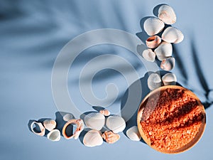 Orange bath salt in a saucer with shells on a blue background with a shadow from a tropical plant. Copyspace, flatlay