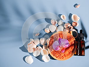 Orange bath salt in a saucer with shells and flower on a blue background with a shadow from a tropical plant. Copyspace