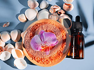 Orange bath salt in a saucer with shells and flower on a blue background with a shadow from a tropical plant. Copyspace