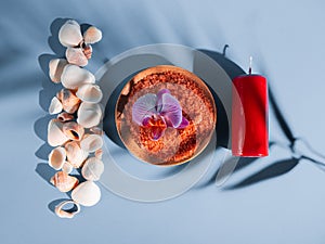 Orange bath salt in a saucer with shells, red candle and flower on a blue background with a shadow from a tropical plant