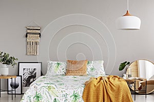 Orange blanket on floral duvet in stylish bedroom interior with handmade macrame, black and white graphics and mirror in wooden