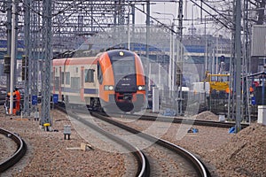 The orange and blue electric train at the railway station. Track, poles and supports for current-carrying lines