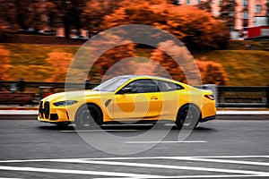 Orange BMW 4 Series coupe G22 is driving along the street with an autumn urban background