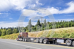 Orange bonnet big rig semi truck tractor transporting two empty flat bed semi trailers driving on the road with forest on the photo
