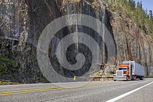 Orange classic bonnet big rig semi-truck tractor transporting cargo in dry van semi trailer driving on the mountain highway road