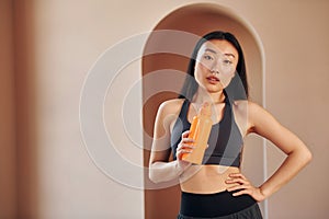 With orange colored bottle. Young serious asian woman standing indoors