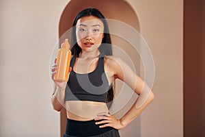 With orange colored bottle. Young serious asian woman standing indoors