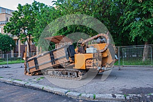 The orange-colored drilling rig stands on the paved city square. In the background there is a fence and green trees