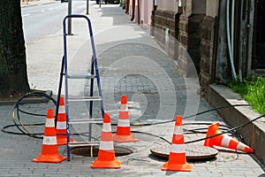 Orange cones installed around the dangerous area on the sidewalk. An open travel hatch. Laying of a new fiber-optic cable in the